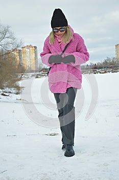 A fashionable girl in a pink fur coat and a black hat walks in the snow in a city park