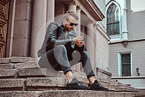 Fashionable guy dressed in a black jacket and jeans using the smartphone sitting on steps against an old building in