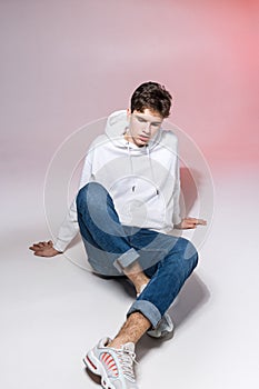 Fashionable handsome young European male model dressed in white sweatshirt, blue jeans and white sneakers posing in studio on a