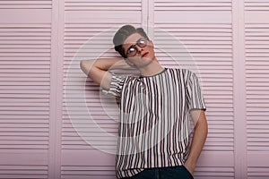 Fashionable handsome young hipster man in stylish glasses with trendy hairstyle in a vintage striped t-shirt posing near a wooden