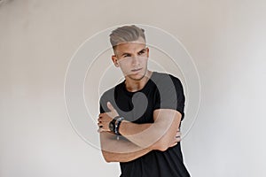 Fashionable handsome young man with hairstyle in a black t-shirt with a watch poses near a white wall on the street