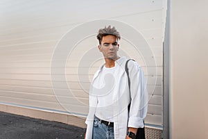 Fashionable handsome young man in stylish clothes with a trendy hairstyle near white wall in the city. European modern guy model