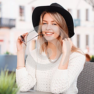 Fashionable happy young hipster woman in a knitted stylish sweater in fashionable sunglasses in an elegant black hat sitting