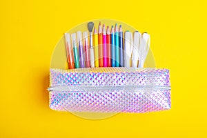 Fashionable sparkling pencil case with multicolored felt-tip pens, pencils, brush and pens on bright yellow background.