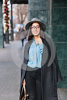 Fashionable joyful young business woman in black glasses walking on street in big city. Blue shirt, grey coat, hat