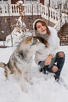 Fashionable joyful young woman having fun with lovely husky dog in snow on the street. True emotions, happy moments in