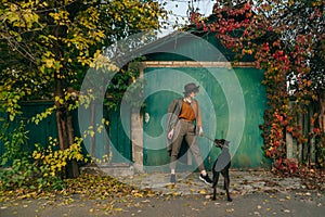 Fashionable lady posing with a dog against the background of a green wall of country cottage and trees with yellow leaves,holding