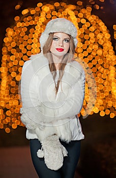 Fashionable lady wearing white fur accessories outdoor with bright Xmas lights in background. Portrait of young beautiful woman