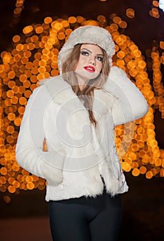 Fashionable lady wearing white fur cap and coat outdoor with bright Xmas lights in background. Portrait of young beautiful woman