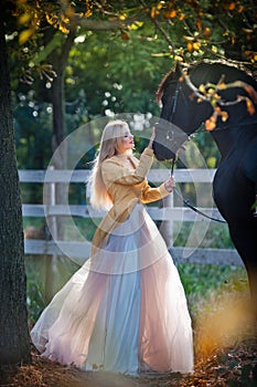 Fashionable lady with white bridal dress near black horse in forest.