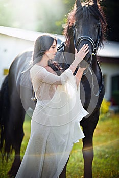 Fashionable lady with white bridal dress near brown horse. Beautiful young woman in a long dress posing with a friendly horse