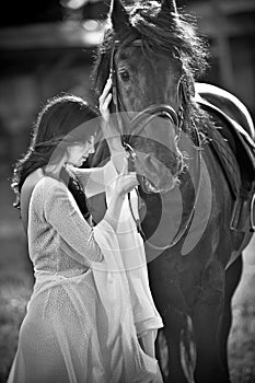 Fashionable lady with white bridal dress near brown horse. Beautiful young woman in a long dress posing with a friendly horse