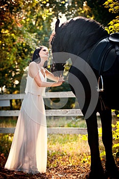 Fashionable lady with white bridal dress near brown horse in nature. Beautiful young woman in a long dress posing with a horse