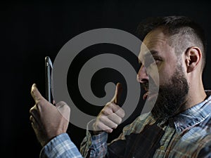 Fashionable man with a beard and mustache grimaces and takes a selfie on the phone on a black background