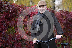 Fashionable man dressed in woolen coat and Irish cap standing with bicycle at a plant fence