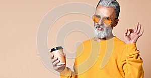 Fashionable man senior looking at camera, having cap with coffee in arms and shows a sign ok with his fingers