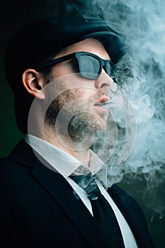 Fashionable man in sunglasses and a leather cap exhales white smoke