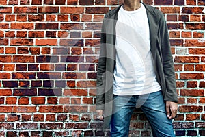 Fashionable man wearing modern stylish casual outfit with grey hoodie, blue jeans and white t-shirt posing near brick wall. Shoppi