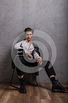 Fashionable model of an American young man in a black leather jacket in a classic white shirt in trendy shoes sits on a chair near