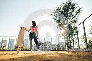 Fashionable model posing with longboard on playground on sunset background. Street stylish photo of girl in casual clothes