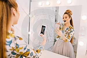 Fashionable model with stylish coiffure, professional makeup making selfie in mirror in hairdresser salon. Expressing