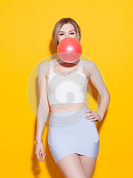 Fashionable modern girl posing in colorful top and skirt inflates the red bubble from chewing gum on yellow background in the stud