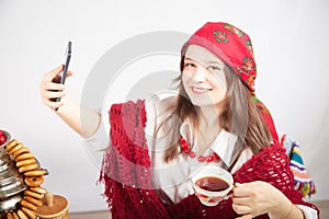 A fashionable modern girl in stylized folk clothes at a table with a samovar, bagels and tea takes a selfie on the