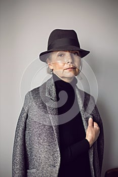 fashionable older woman in black clothes and a hat stands against a white wall