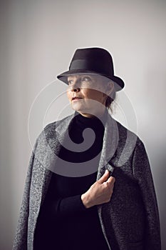 fashionable older woman in black clothes and a hat stands against a white wall