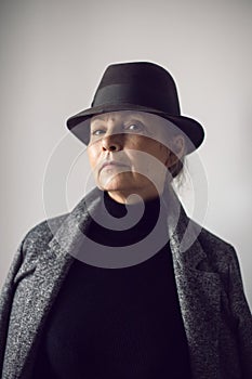 fashionable older woman in black clothes and a hat stands against a white wall