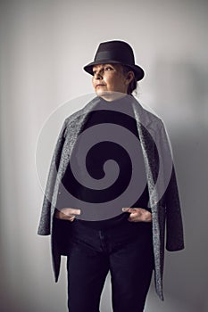 fashionable older woman in black clothes and a hat stands against a white wall
