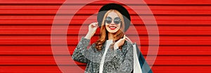 Fashionable portrait beautiful happy smiling woman with shopping bags wearing gray coat, hat on colorful red background