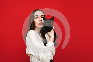 Fashionable portrait of a beautiful woman in a white shirt stands on a red background with a black cat in hand, looks into the