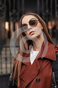 Fashionable portrait beautiful young hipster woman in trendy trench coat in stylish dark sunglasses with long hair near vintage