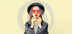 Fashionable portrait of stylish young woman covering her eyes with red heart shaped lollipop blowing her lips sending sweet kiss