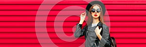 Fashionable portrait of stylish young woman posing wearing black rock biker jacket, round hat and backpack on red background,