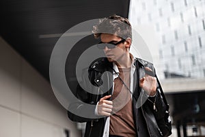 Fashionable portrait of a young man hipster in stylish dark sunglasses with a hairstyle in trendy black leather jacket in the