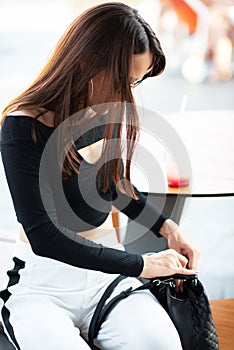 Fashionable pretty stylish girl in outdoor cafe open her bag. Beautiful young brunette woman wearing white trousers and black