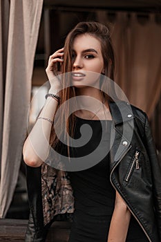 Fashionable pretty young woman model in a vintage leather black jacket in a black stylish dress straightens hair indoors.