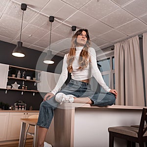 Fashionable pretty young woman in a white stylish top in trendy blue jeans resting on the kitchen vintage wooden table indoors.