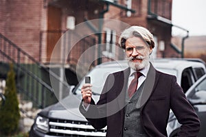 Fashionable senior man with gray hair and beard standing outdoors on the street near his car with keys in hand