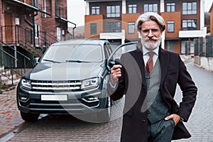 Fashionable senior man with gray hair and beard standing outdoors on the street near his car with keys in hand