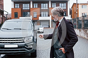 Fashionable senior man with gray hair and beard standing outdoors on the street near his car with keys in hand