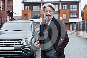 Fashionable senior man with gray hair and beard standing outdoors on the street near his car with keys in hand