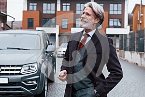 Fashionable senior man with gray hair and beard standing outdoors on the street near his car with keys in hand