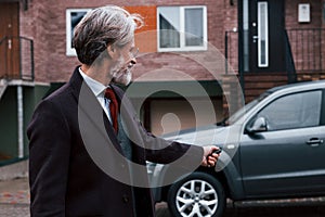 Fashionable senior man with gray hair and beard standing outdoors on the street near his car with keys in hand