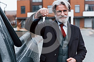 Fashionable senior man with gray hair and beard standing outdoors on the street near his car with keys in hand