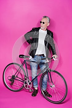 Fashionable senior man wearing leather jacket and sunglesses standing with bicycle