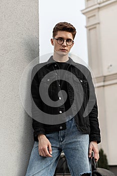 Fashionable serious young man fashion model in stylish jeans youth casual wear in vintage glasses with backpack resting near wall