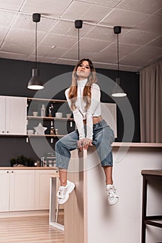 Fashionable sexy young woman model in a fashionable white top in blue vintage jeans in trendy sneakers sits on the kitchen table